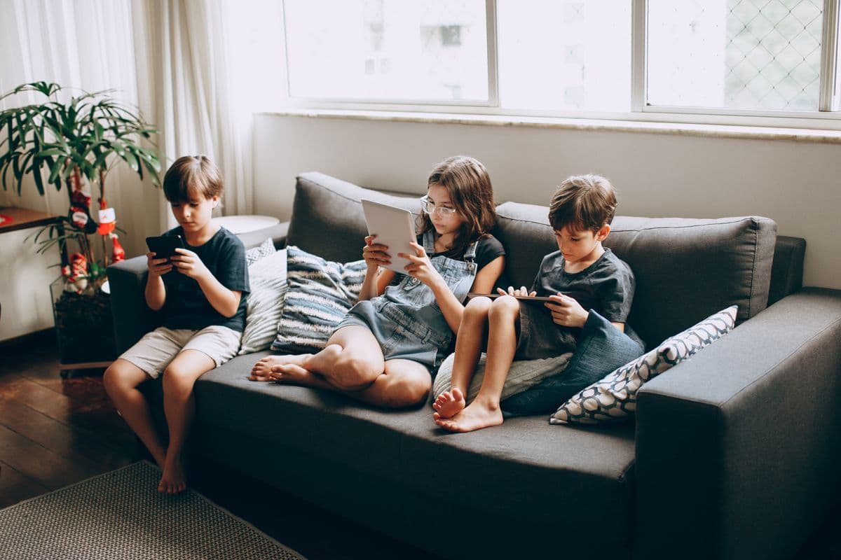 Three children sitting together on a grey sofa by a bright window, each looking at their own phone or tablet